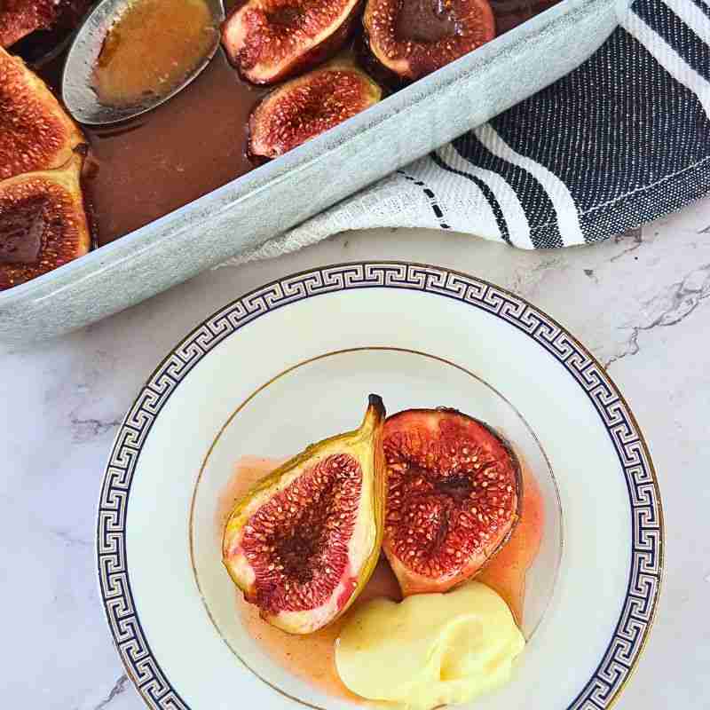 honey and orange baked figs with orange mascarpone cream on white and gold plate with kitchen towel and baking dish of figs